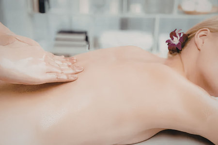 a woman lying on her stomach with her face turned away, receiving a massage on her back from a therapist's hand, in a spa setting with blurred backgroundの写真素材