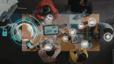 Professionals collaborate around a table with laptops and digital interfaces displaying data...