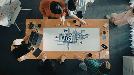 Group of professionals gathered around a wooden table with laptops and a whiteboard discussing...