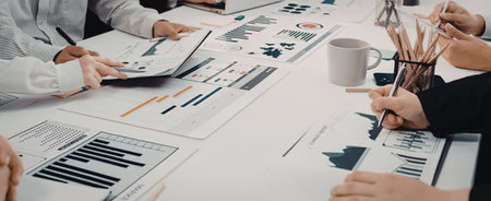 Business professionals collaborate around a white table analyzing data and charts with pens and...
