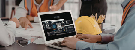 A construction team gathered around a table reviewing blueprints on a laptop computer. The team...