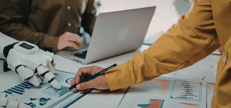 Human worker in orange shirt collaborates with robotic technology to analyze data on graphs and...