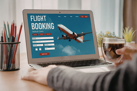 Person books flight online using laptop while holding a cup of coffee on a wooden desk with...