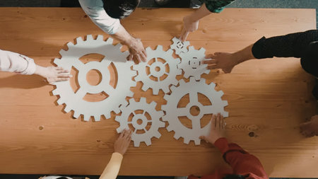 People put together interlocking white gear puzzle pieces on a wooden table, symbolizing teamwork...
