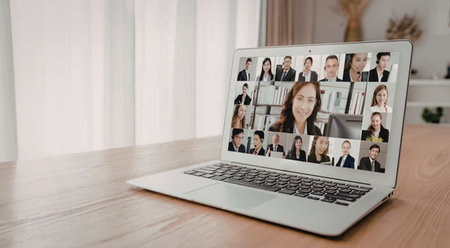 A laptop on a wooden table displays a virtual meeting with multiple participants on the screen.