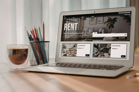A laptop on a wooden desk displaying a rental website with a cup of coffee and colored pencils nearby.