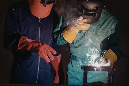 Two workers wearing protective gear are engaged in a welding process, with one actively welding and the other observing.