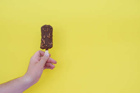 Hand of man holds chocolate ice cream on a stick, as eskimo, on a yellow surface background.の写真素材