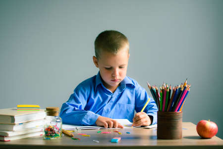 Boy in a blue shirt sitting at the table and draws in a notebook, on the table books, pencils, Apple and other stationery. Boy learning.の写真素材