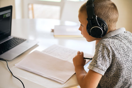 School boy in headphones writing in notebook and using laptop during online lesson. Online education...