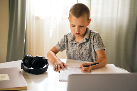 School boy writing in notebook and using laptop while doing schoolwork at home or during online lesson. Online education concept.の写真素材