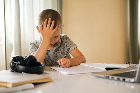 School boy writing in notebook and using laptop while doing schoolwork at home or during online lesson. Online education concept.の写真素材