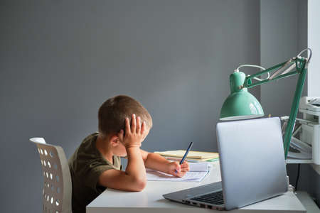 The schoolboy sitting at the table and using laptop while doing schoolwork at home. Homeschooling...