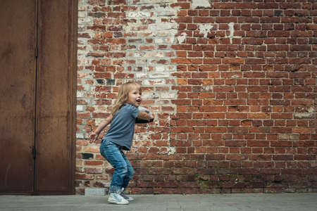 Little cute girl in a blue t-shirt and jeans with a cap is dancing on the background of an old brick wall.の写真素材