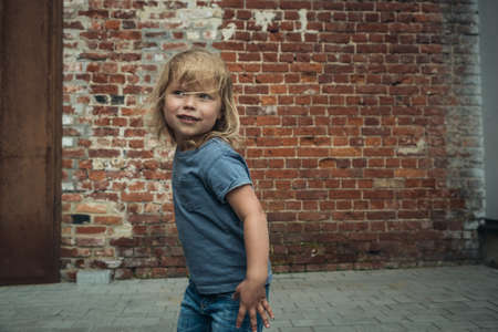 Little cute girl in a blue t-shirt and jeans with a cap is dancing on the background of an old brick wall.の写真素材