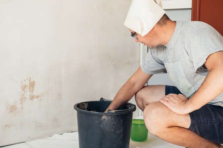 A man in a paper cap makes repairs at home, prepares a wall for pasting wallpaper. Renovation diy new home painting wall.の写真素材