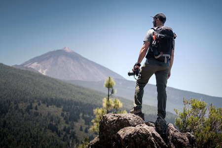 A photographer observes the horizon with his camera in hand from the top of a mountain. You see the landscape with forests, mountains and a volcano. Travel or adventure concept.の写真素材