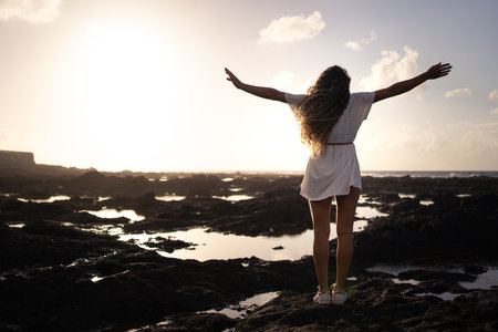 A blonde woman with curly hair and a white dress standing with her arms wide open looking out to sea with a sky full of clouds and colors at sunset. Concept peace, relaxation, tranquility, happinessの写真素材
