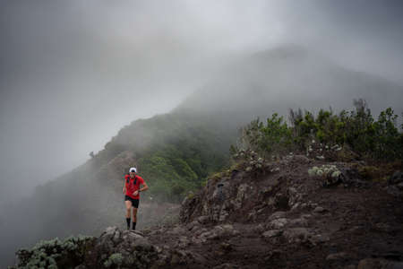 A man in a cap and red shirt doing trail running through the mountain and forest with fog and cloudy weather.の写真素材