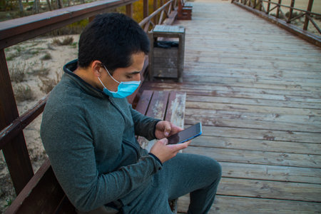 Man sitting on a wooden bench at the beach with a covid mask consulting his mobile phone