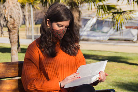 Woman with surgical mask playing guitar sitting on a park bench.の写真素材