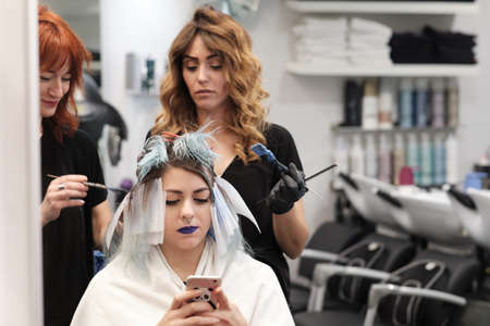 two young hairdresser dyeing hair of female client while she looks to her mobileの写真素材