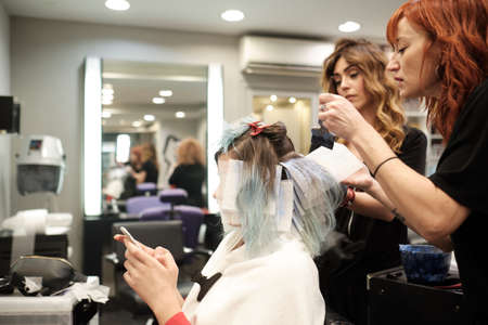 two young hairdresser dyeing hair of female client while she reads her mobile phoneの写真素材