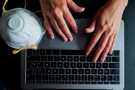 a closeup image of a woman working and typing on laptop computer keyboard with a mask during coronavirus crisisの写真素材
