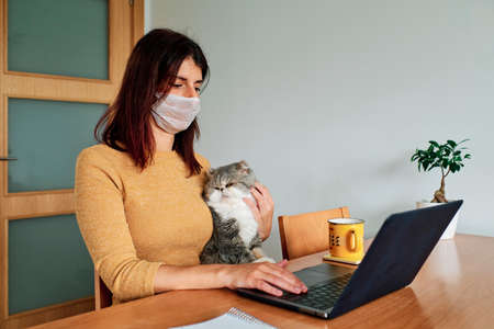 Coronavirus. A woman in quarantine for coronavirus with her cat wearing protective mask and working from home using laptop. Video call. Video conference. Stay home conceptの写真素材