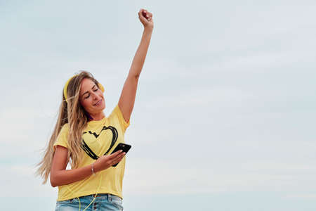 young blonde woman using yellow headphones in front of the beach on a summer cloudy dayの写真素材