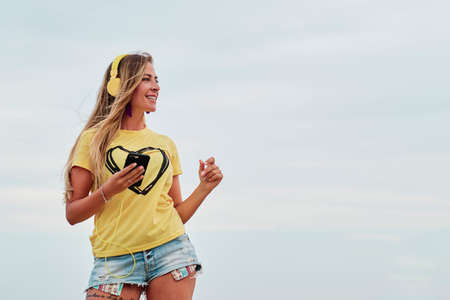young blonde woman using yellow headphones in front of the beach on a summer cloudy dayの写真素材