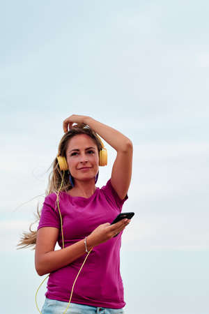 young woman with sky behind listening to headphones with her mobile phone