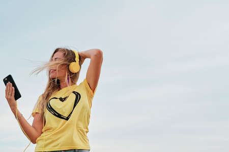 young blonde woman using yellow headphones in front of the beach on a summer cloudy day