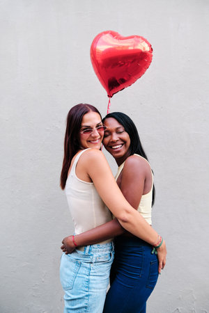 Cheerful lesbian couple hugging while holding a red heart-shaped balloon.の写真素材