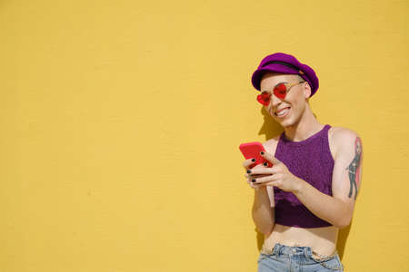 Happy non-binary person smiling while using a mobile phone outdoors against a yellow wall.の写真素材