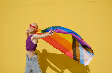 Young stylish non-binary person carrying the lgbt rainbow flag against a yellow wall.