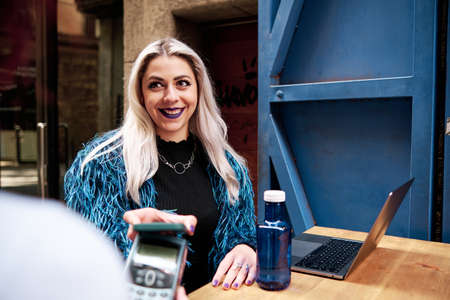 Alternative woman smiling while paying contactless with her mobile phone at a coffee shop.