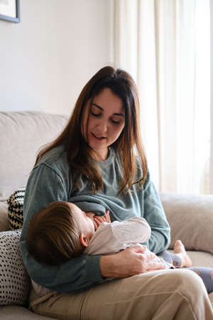 Mother breastfeeding her daughter while sitting on the sofa at home.の写真素材