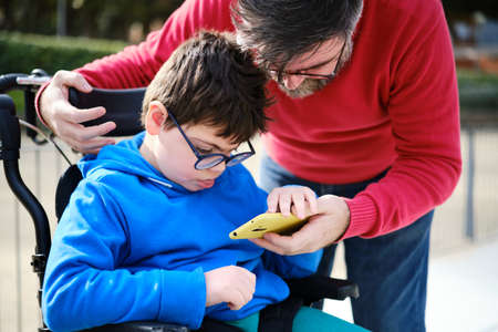 Disabled child on wheelchair and his father using a mobile phone together during a walk outdoors.