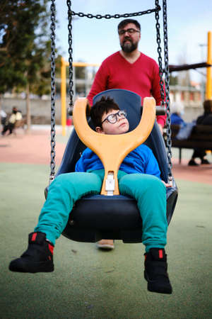 Disabled boy swinging on a special needs swing while enjoying with his father at the playground.の写真素材