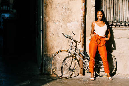 Young African American woman smiling while standing outdoors on the street with her bicycle.の写真素材