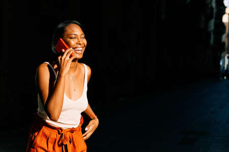 Smiling African American woman talking on the phone while being lit by sunlight outdoors.の写真素材