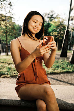 Young Asian woman smiling while using her mobile phone sitting on a bench in the park.の写真素材