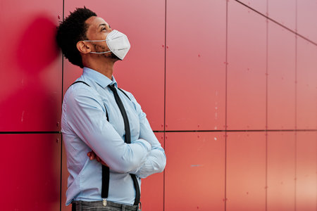 Businessman with a face mask looking up while leaning on a wall with his arms crossed.の写真素材