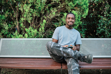 Man looking at camera and smiling while using mobile phone sitting on a bench outdoors.の写真素材