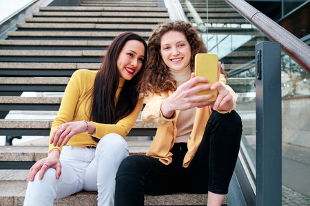 Two women smiling while taking selfies with a mobile phone sitting on stairs outdoors.