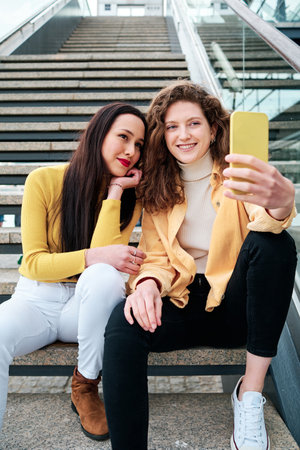 Two female friends smiling while taking selfies with a mobile phone sitting on stairs outdoors.の写真素材