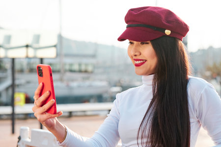 Stylish woman with beret smiling while taking selfies with mobile phone outdoors.