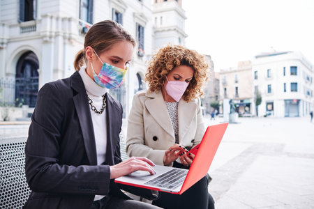 Two businesswoman with face masks using mobile phone and laptop while sitting outdoors.の写真素材