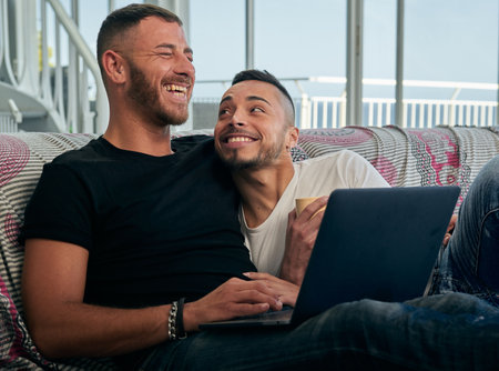 Male couple smiling and having fun together while using a laptop on the sofa.の写真素材